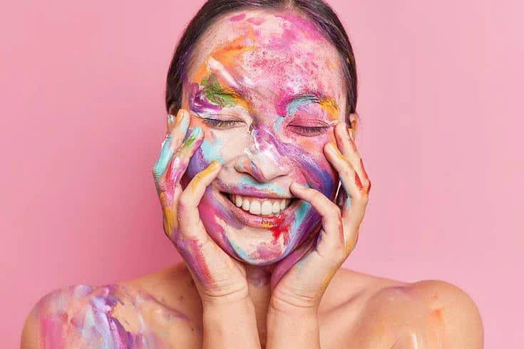 A person with paint on their hands and shoulders is holding their face, set against a vibrant pink background.