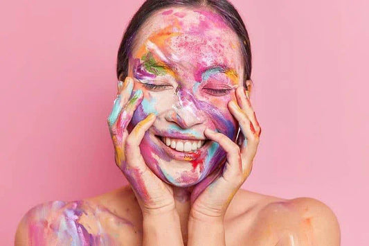 A person with paint on their hands and shoulders is holding their face, set against a vibrant pink background.