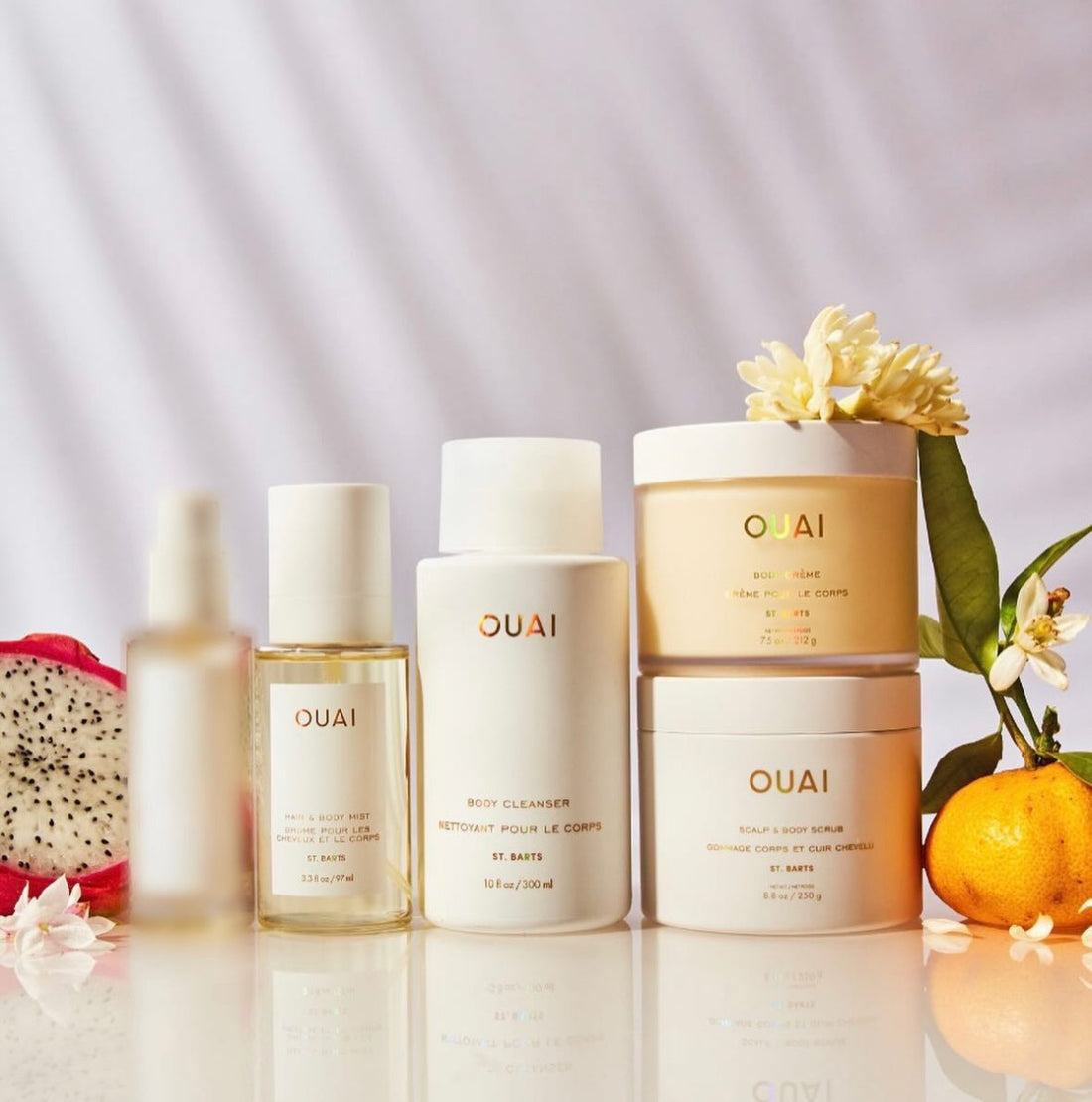 A collection of OUAI body care products, including a body cleanser, body cream, mist, and scalp scrub, arranged with fruit and flowers.
