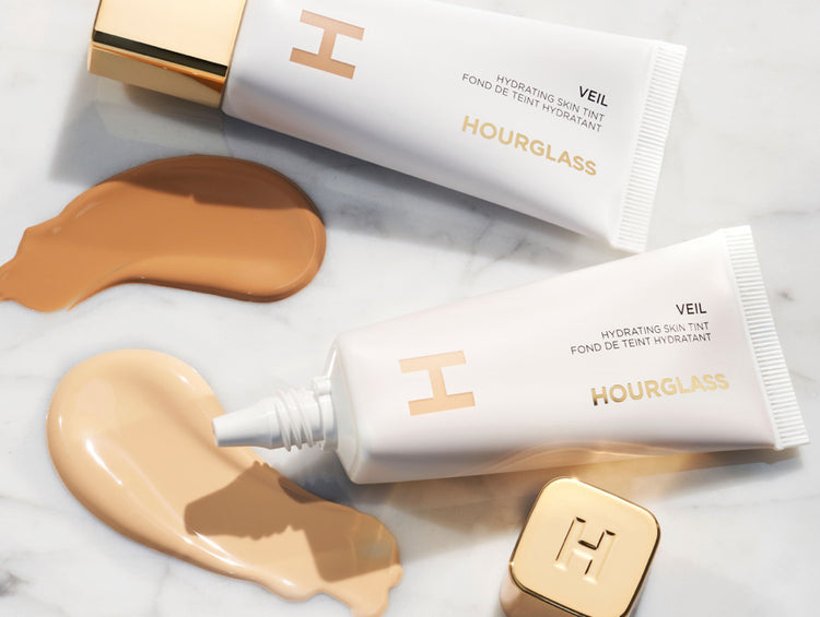 Two tubes of Hourglass Veil Hydrating Skin Tint on a marble surface, accompanied by swatches of creamy foundation shades.