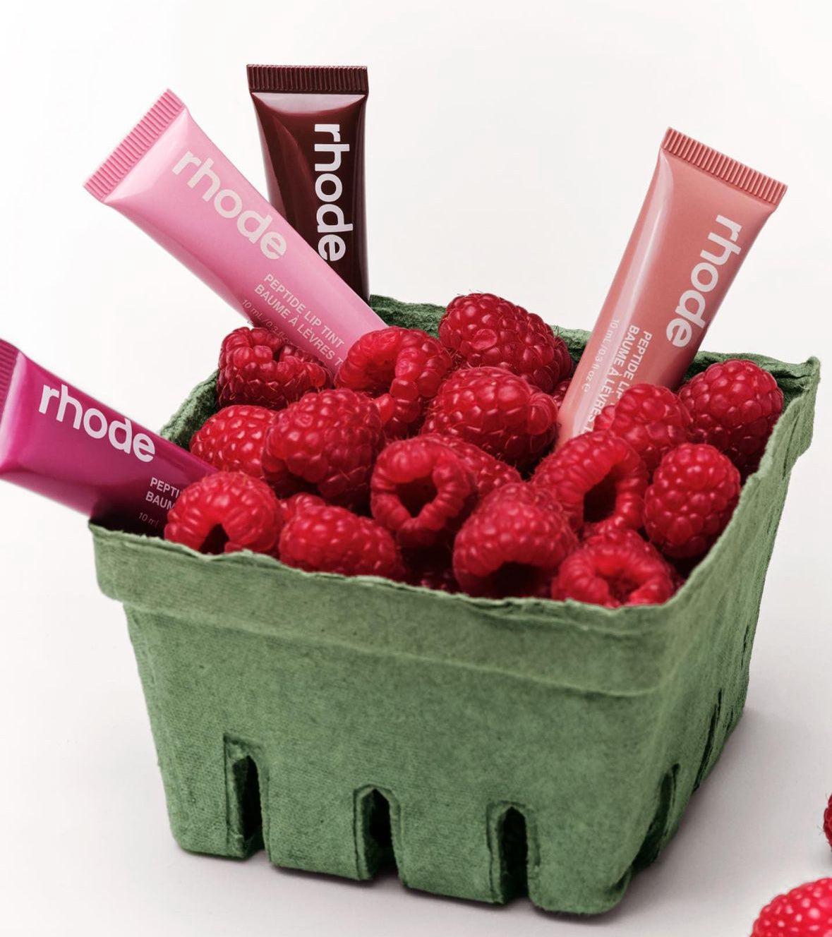 A green basket filled with fresh raspberries and vibrant rhode peptide lip tint tubes in pink and burgundy shades.