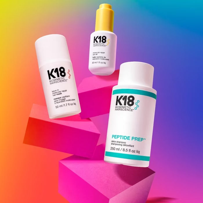 Three K18 hair care products in vibrant packaging on colorful geometric shapes, featuring a repair mask, treatment oil, and detox shampoo.