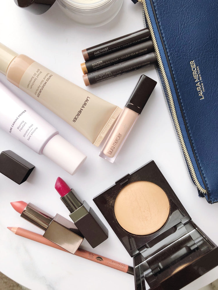 Laura Mercier cosmetics arranged on a white marble surface, including primer, tinted moisturizer, concealer, eye crayons, lipstick, lip liner, and powder. A blue makeup bag bearing the logo is positioned at the top right.