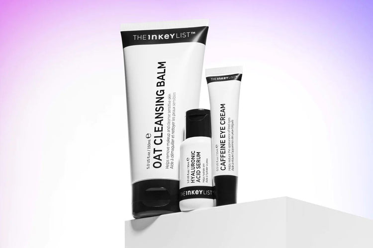 Three skincare products by The Inkey List: Oat Cleansing Balm, Hyaluronic Serum, and Caffeine Eye Cream on a white pedestal.