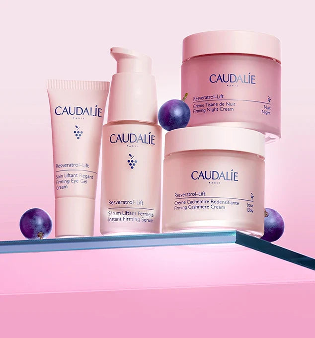 Four Caudalie skincare products from the Resveratrol-Lift line, displayed on a glass shelf against a pink background, featuring grapes.