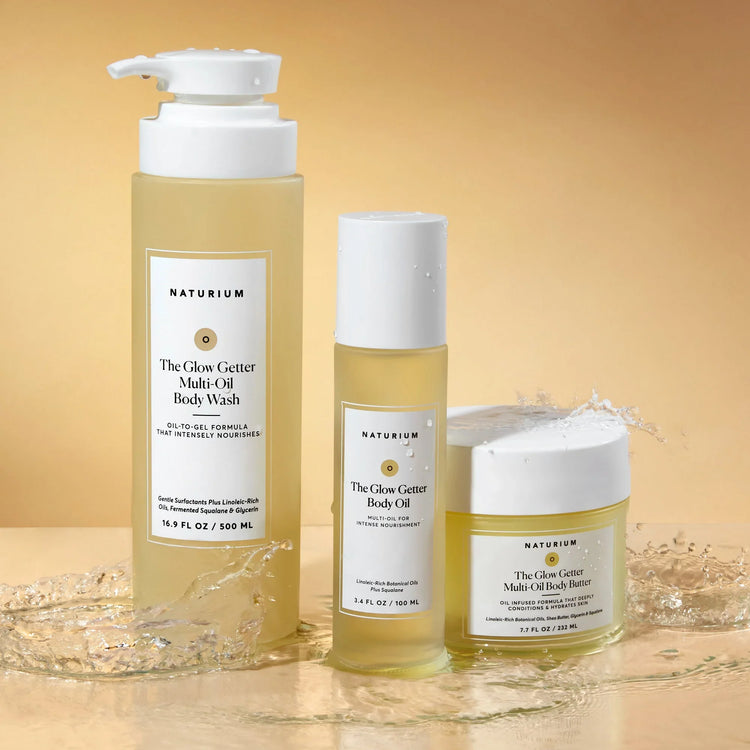 Three Naturium Glow Getter body products on a beige background: a body wash, body oil, and body butter, with splashes of water.