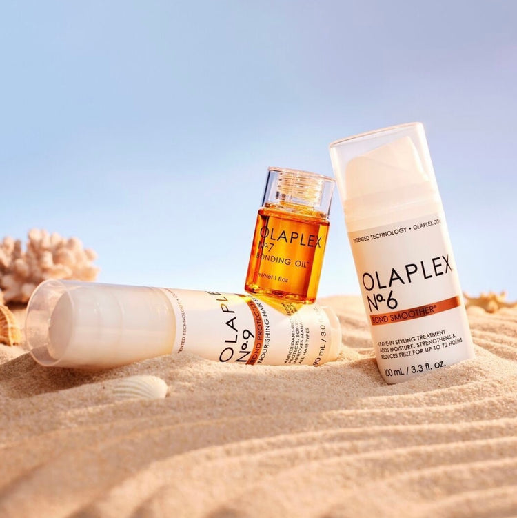 Three Olaplex hair care products arranged on sandy beach: No. 9 Nourishing Hair Serum, No. 7 Bonding Oil, and No. 6 Bond Smoother.