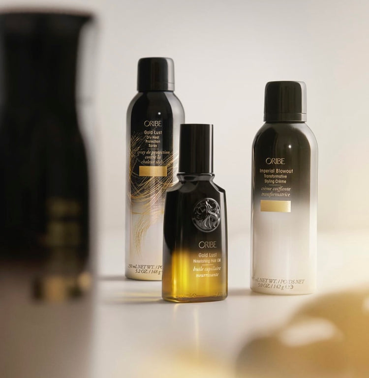 Three hair-care bottles of varying sizes by Oribe, including spray, crème, and oil products.