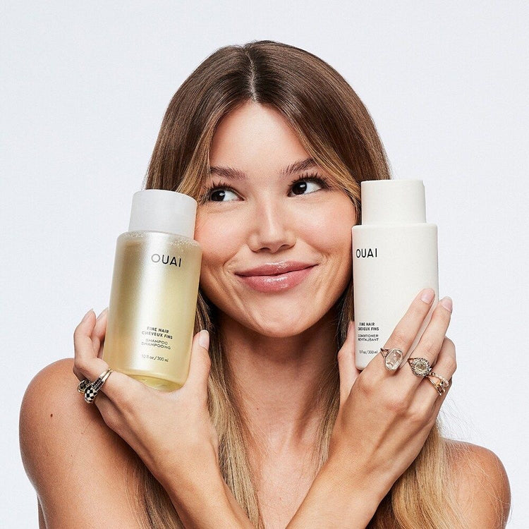 A person holds two OUAI hair products, a yellow shampoo and a white conditioner, showcasing a stylish, minimalist look.