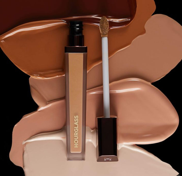 Hourglass concealer tube beside swatches of various skin tones on a black background, showcasing its versatility in complexion.
