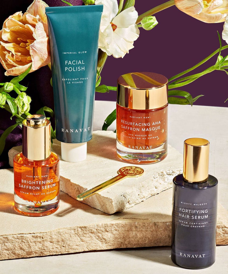 A collection of Ranavat beauty products including serums and a facial polish, with flowers and a decorative spoon on a stone surface.