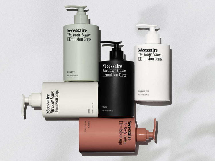 Five bottles of Nécessaire body lotion in various colors—fragrance-free, eucalyptus, sandalwood, bergamot, and a pink shade—arranged artistically.