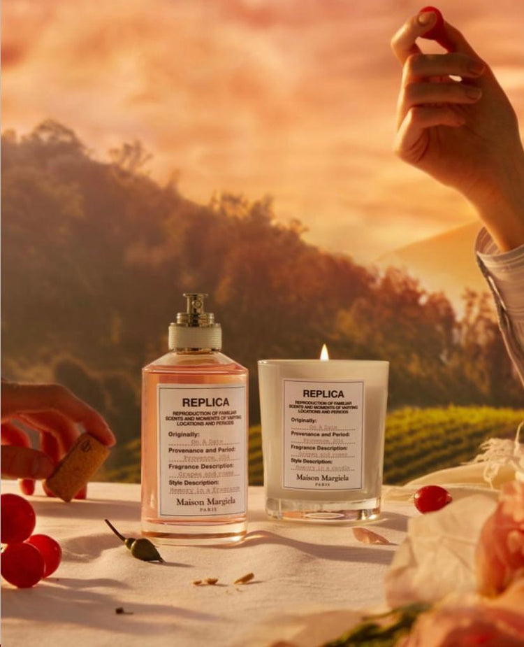 Two Maison Margiela Replica fragrance products on a table with grapes, set against a scenic sunset vineyard backdrop.