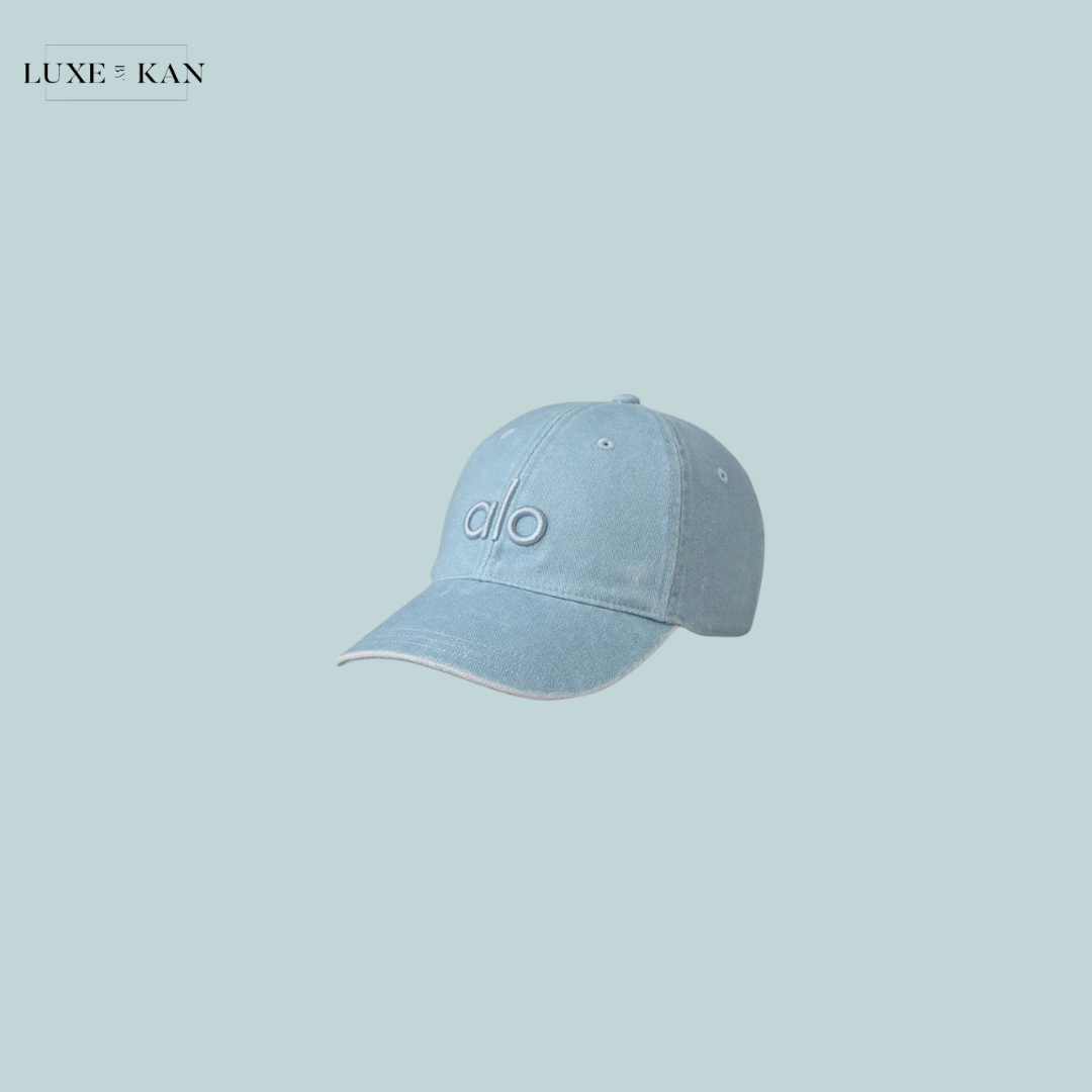 Light blue baseball cap with embroidered "alo" logo, set against a soft pastel background. Stylish and casual accessory.