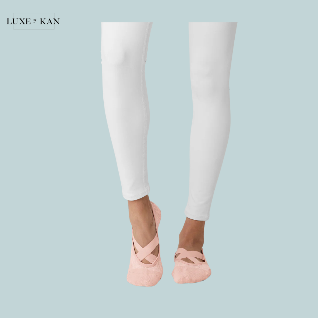 A pair of feet in light pink ballet slippers, wearing white fitted pants against a soft blue background. Featuring a Luxe & Kan logo.