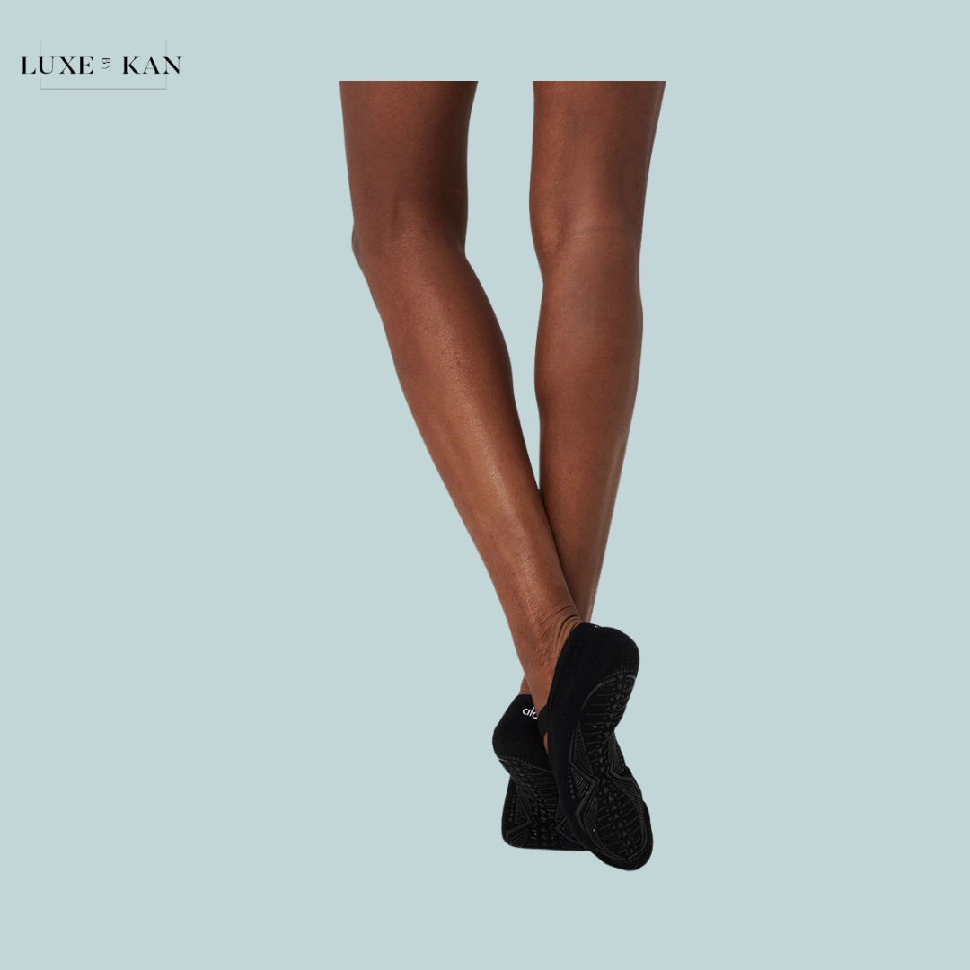 A pair of crossed brown legs in black slip-on shoes, set against a light blue background. The focus is on the legs and footwear.