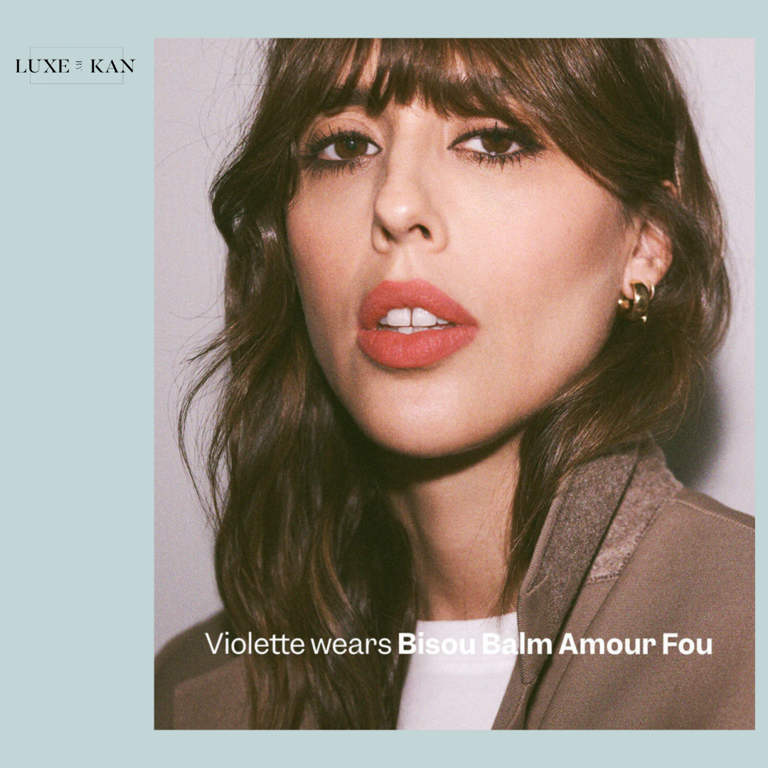 A woman with wavy hair wears a stylish brown jacket, showcasing Bisou Balm Amour Fou products at the bottom of the image.