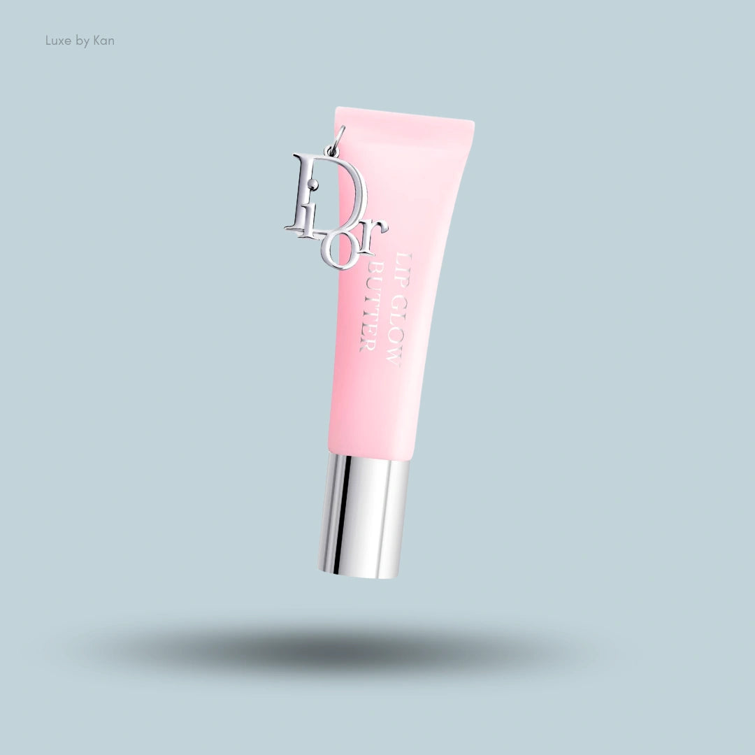 A tube of Dior Lip Glow Butter in pastel pink with silver accents, featuring a silver Dior logo charm hanging from the tube.
