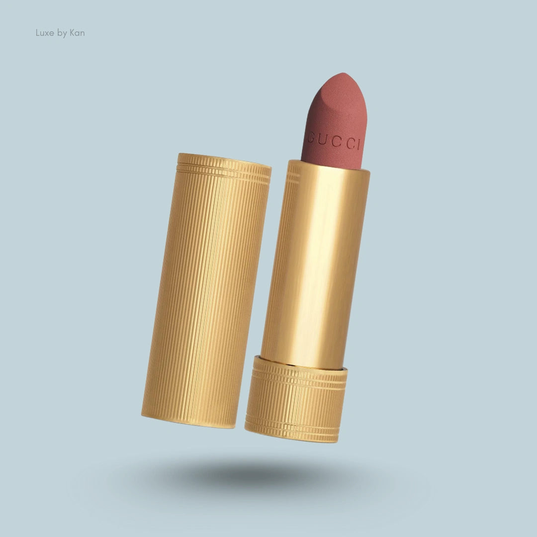 Gucci Rouge à Lèvres Mat lipstick with gold ribbed casing and peggy taupe colored lipstick on a light blue background.