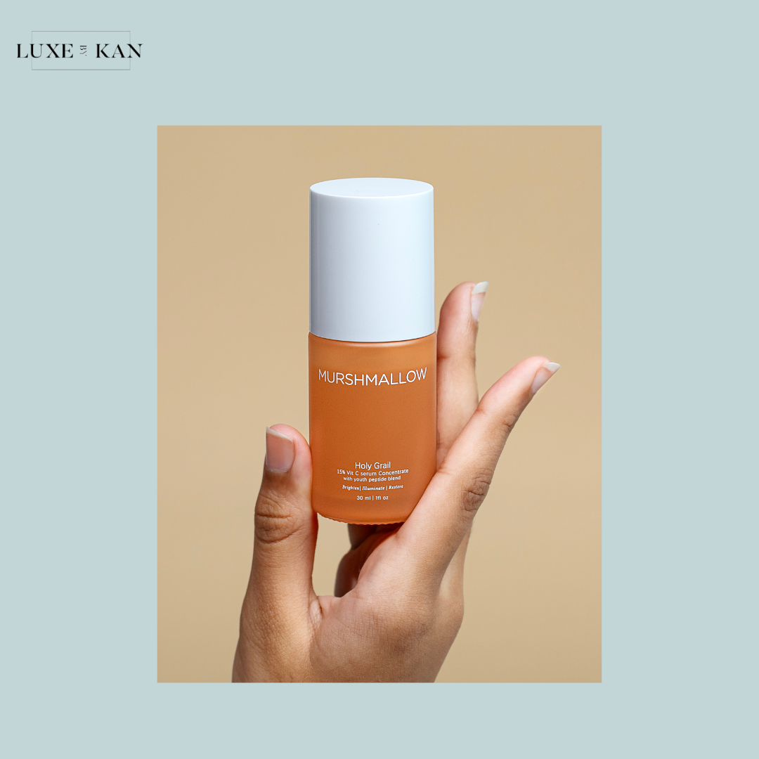 A hand holds a bottle of Murshmallow "Holy Grail" serum against a soft beige background, showcasing its sleek design.