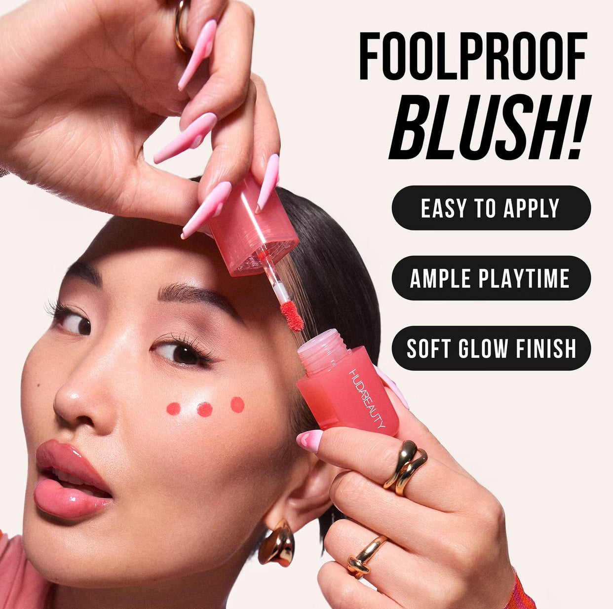 A hand with long pink nails holds a tube of Huda Beauty blush, showcasing its easy application and glowing finish.