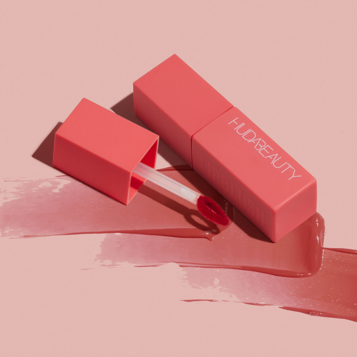 A pink Huda Beauty creamy lip and cheek stain tube and applicator resting on a rosy surface, showcasing its vibrant color and sleek packaging.