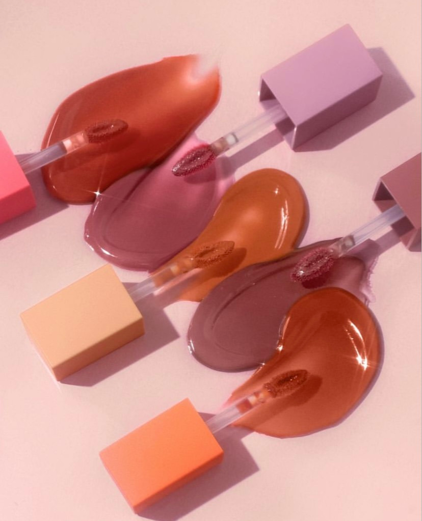 An arrangement of huda beauty liquid lipsticks in various shades of pink, nude, and coral, with matching square caps on a soft pink background.