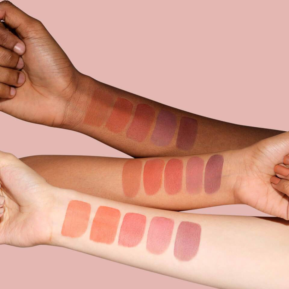 Three arms display a variety of makeup swatches in warm and cool tones against a soft pink background.