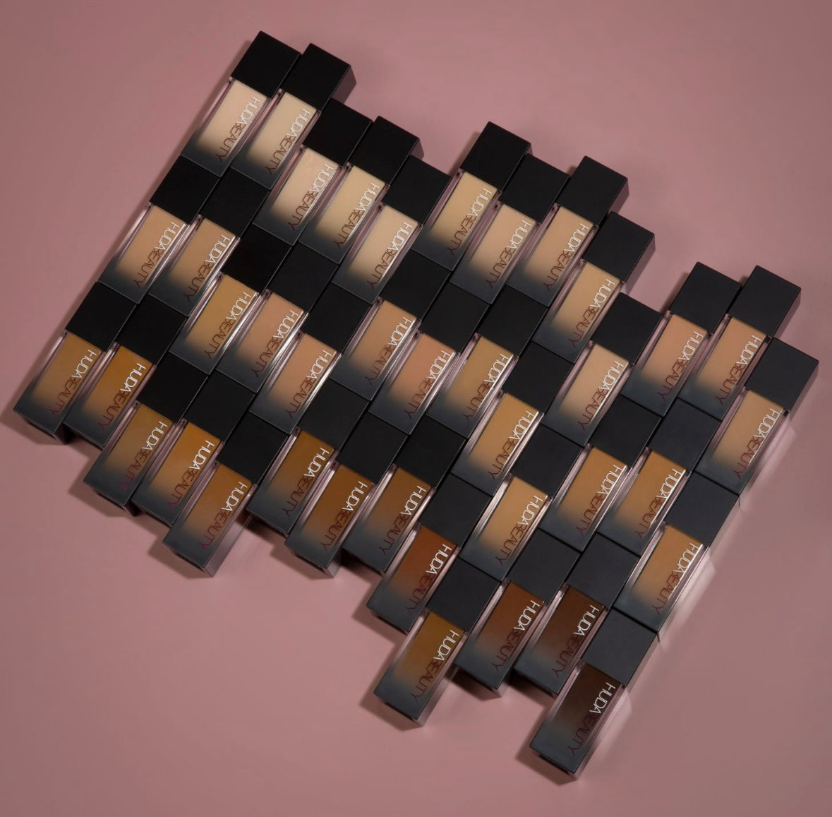 A neatly arranged display of Huda Beauty fauxfilter foundation bottles on a soft pink surface, showcasing various shades.