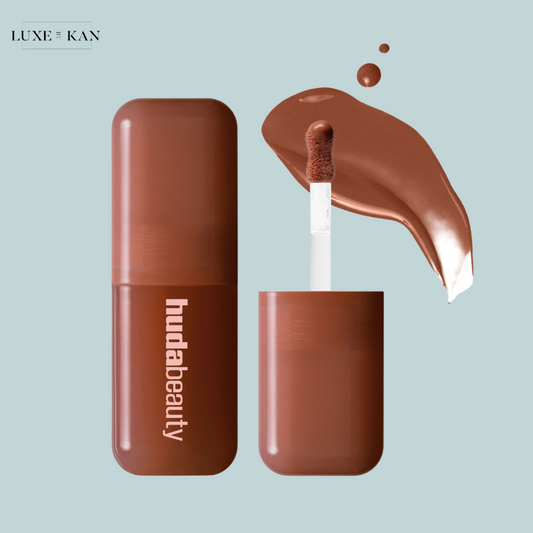 A brown lip product from Huda Beauty, featuring a wand applicator and a swatch of rich chocolate color on a soft blue background.