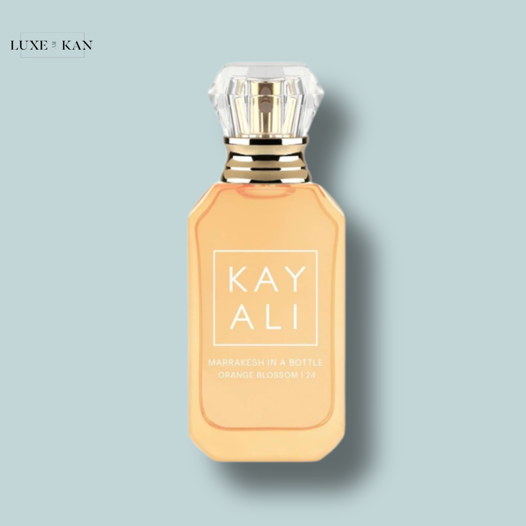 A stylish bottle of Kayali's "Marrakesh in a Bottle" perfume, featuring a golden-orange hue and elegant design against a soft blue background.