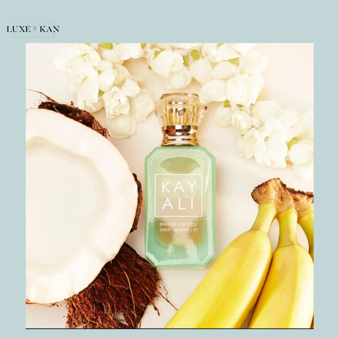 A bottle of KAYALI Sweet Vanilla perfume surrounded by coconut, bananas, and white flowers on a soft background.