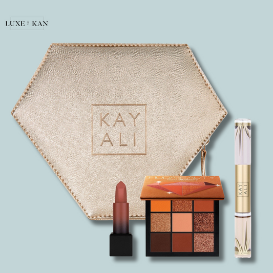 A hexagonal, gold makeup pouch containing a mauve lipstick, an eyeshadow palette with warm tones, and a clear lip gloss, all branded Kayali.