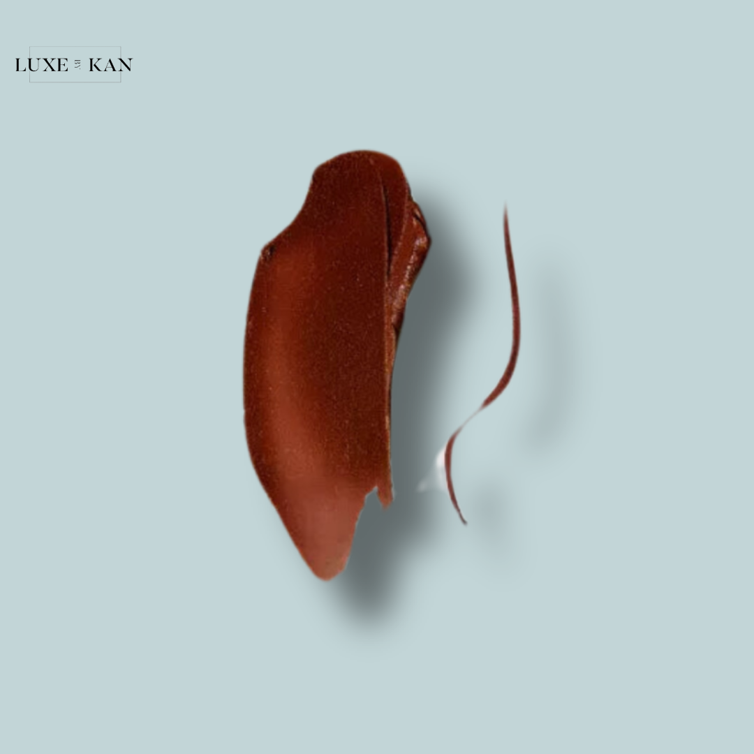 A rich, dark brown lip product swatch against a soft blue background, showcasing its smooth texture and depth. Logo in the top left corner.