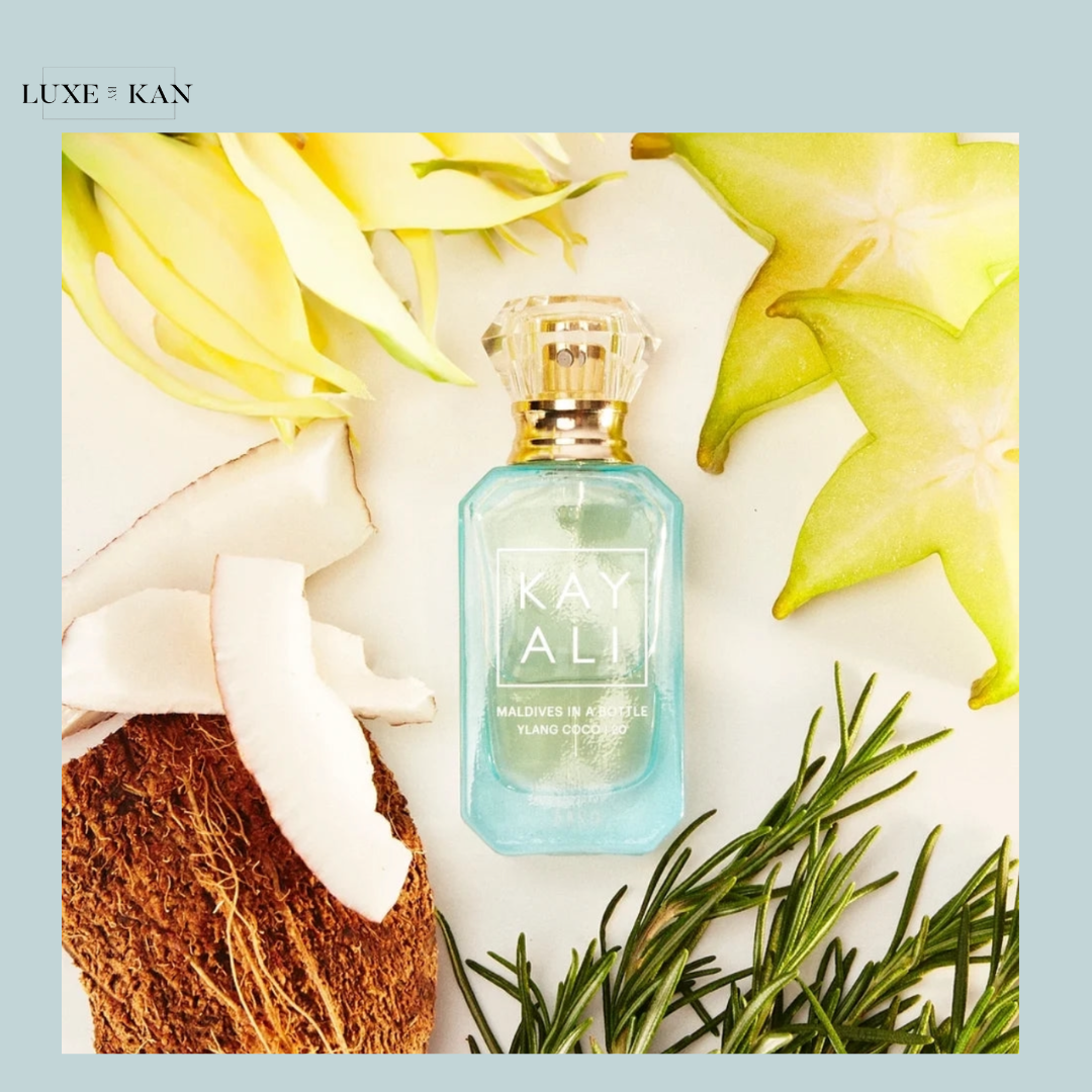 A turquoise perfume bottle labeled "KAYALI Maldives in a Bottle" surrounded by coconut, yellow flowers, and green leaves.