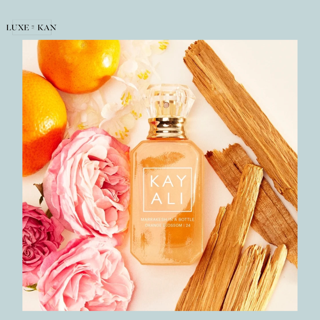 A bottle of Kayali Marrakesh perfume surrounded by pink roses, oranges, and wooden elements on a light background.