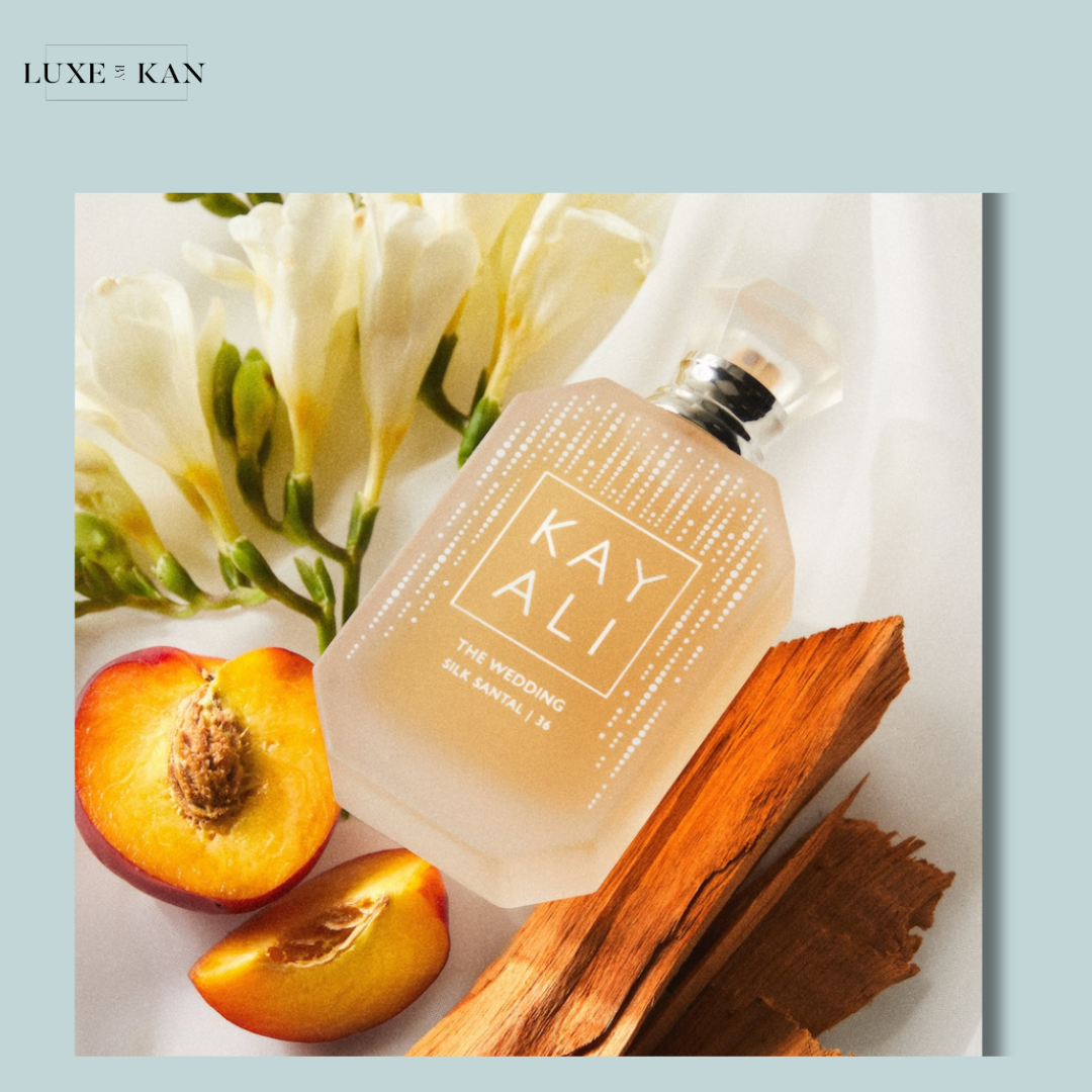 A bottle of KAYALI perfume, "THE WEDDING," surrounded by peach slices, wood accents, and white flowers on a soft backdrop.