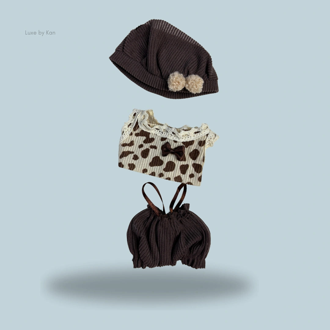 Labubu cocoa cuddle 3-piece set includes a cocoa-colored knit hat with pom poms, a cow-print top with lace trim and a brown bow, and cocoa-colored bloomers.