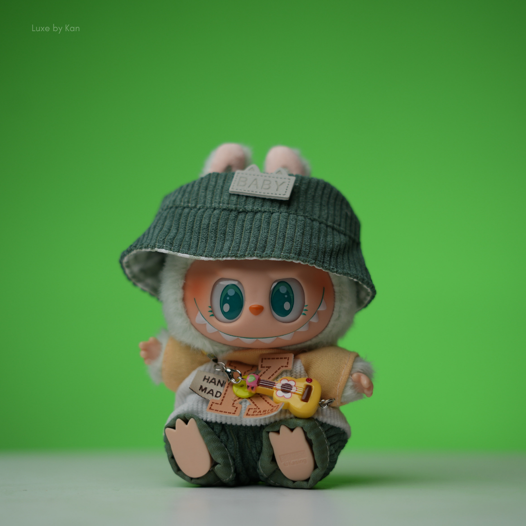 The Labubu doll is dressed in a little explorer three-piece set, complete with a green corduroy bucket hat, a fluffy green-and-white outfit, and a tiny yellow ukulele.