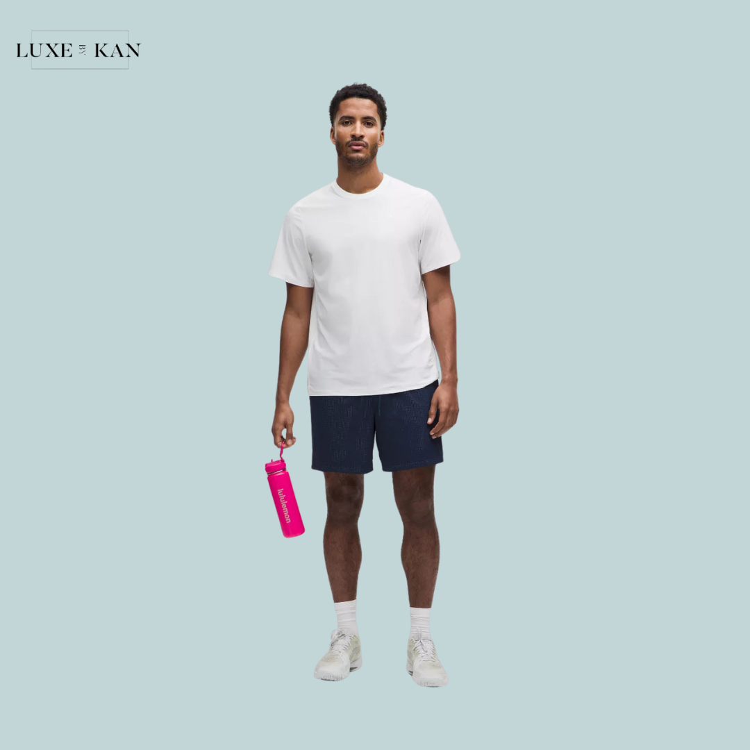 A male model stands against a light blue background, wearing a white T-shirt and navy shorts, holding a bright pink Back to Life Sport Bottle with a straw lid.