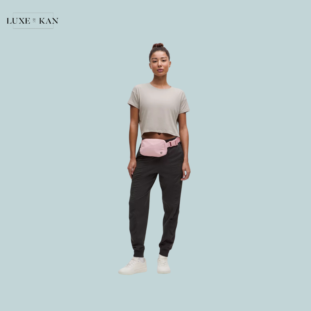 A person wears a light gray cropped t-shirt, black joggers, and a pink lululemon everywhere belt bag.