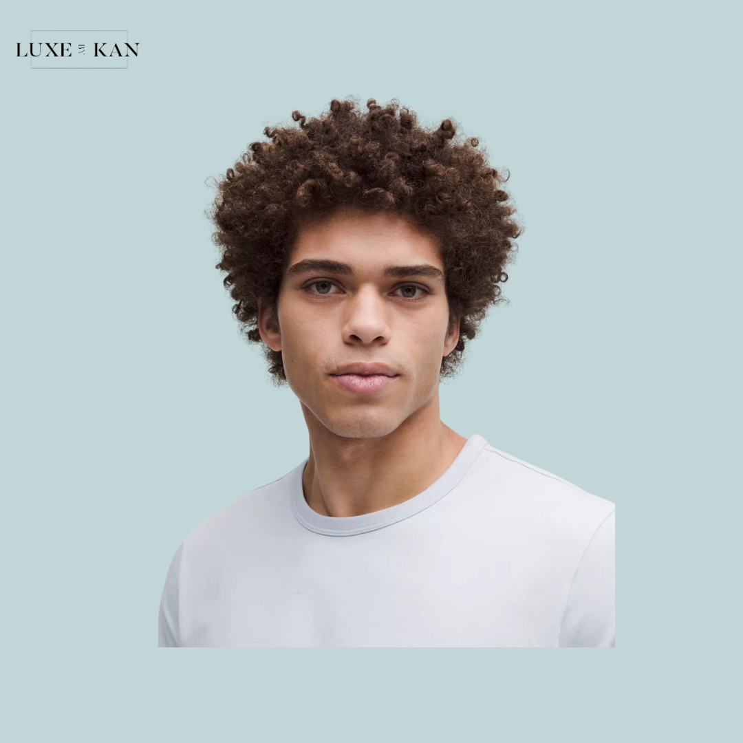 A person with curly hair wearing a light gray t-shirt poses against a soft blue background, highlighted by the brand logo Luxe & Kan.