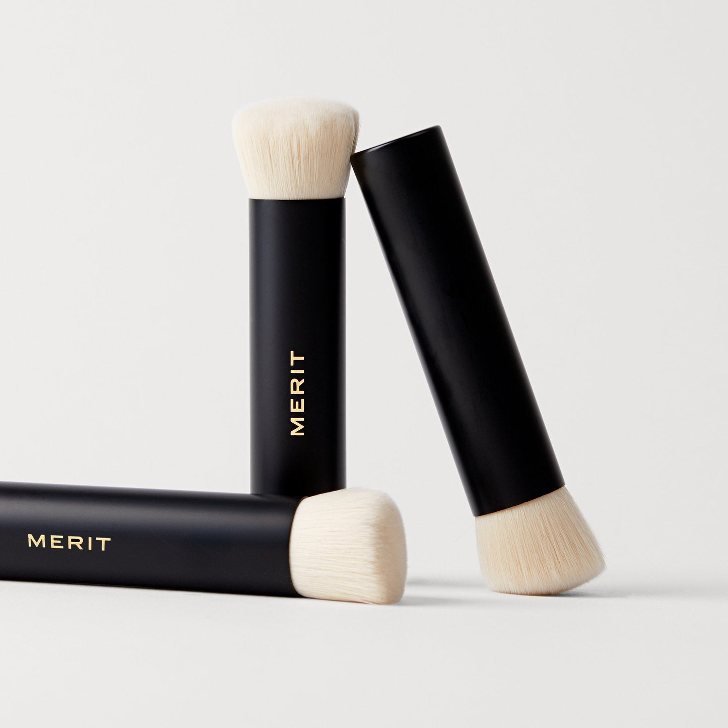 Three black makeup brushes with soft white bristles, featuring the gold "MERIT" logo on the handles, arranged in a casual position.