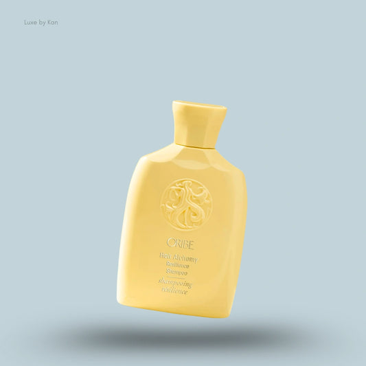 Oribe Hair Alchemy Resilience Shampoo in a yellow bottle with the brand's name and logo against a light blue background.