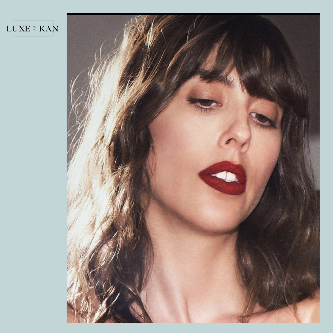 A woman with wavy hair is partially lit, and the red color of Petal Bouche matte lipstick is visible on her lips.