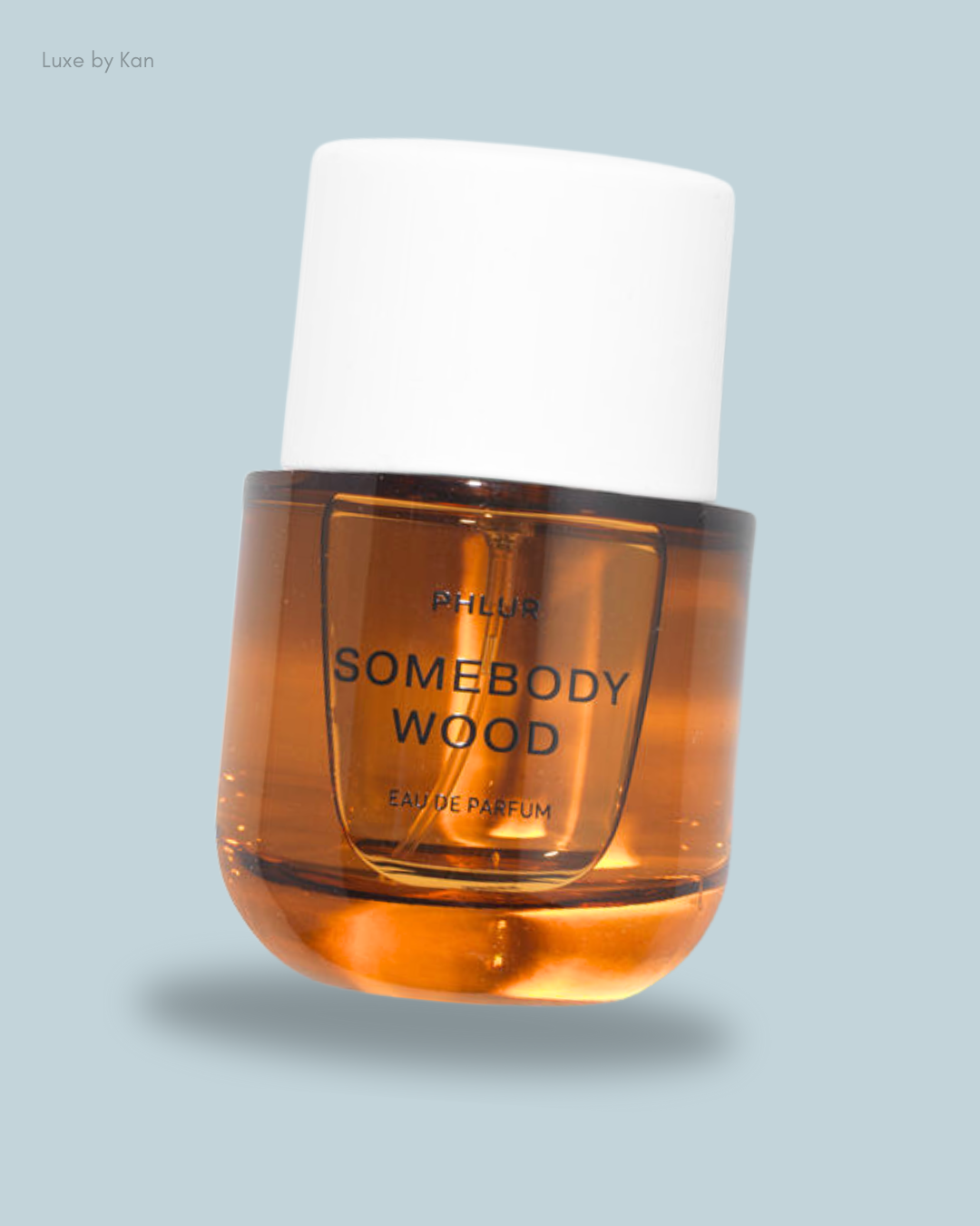 A bottle of Phlur's "Somebody Wood" eau de parfum, featuring a transparent amber liquid and a white cap against a soft gray backdrop.