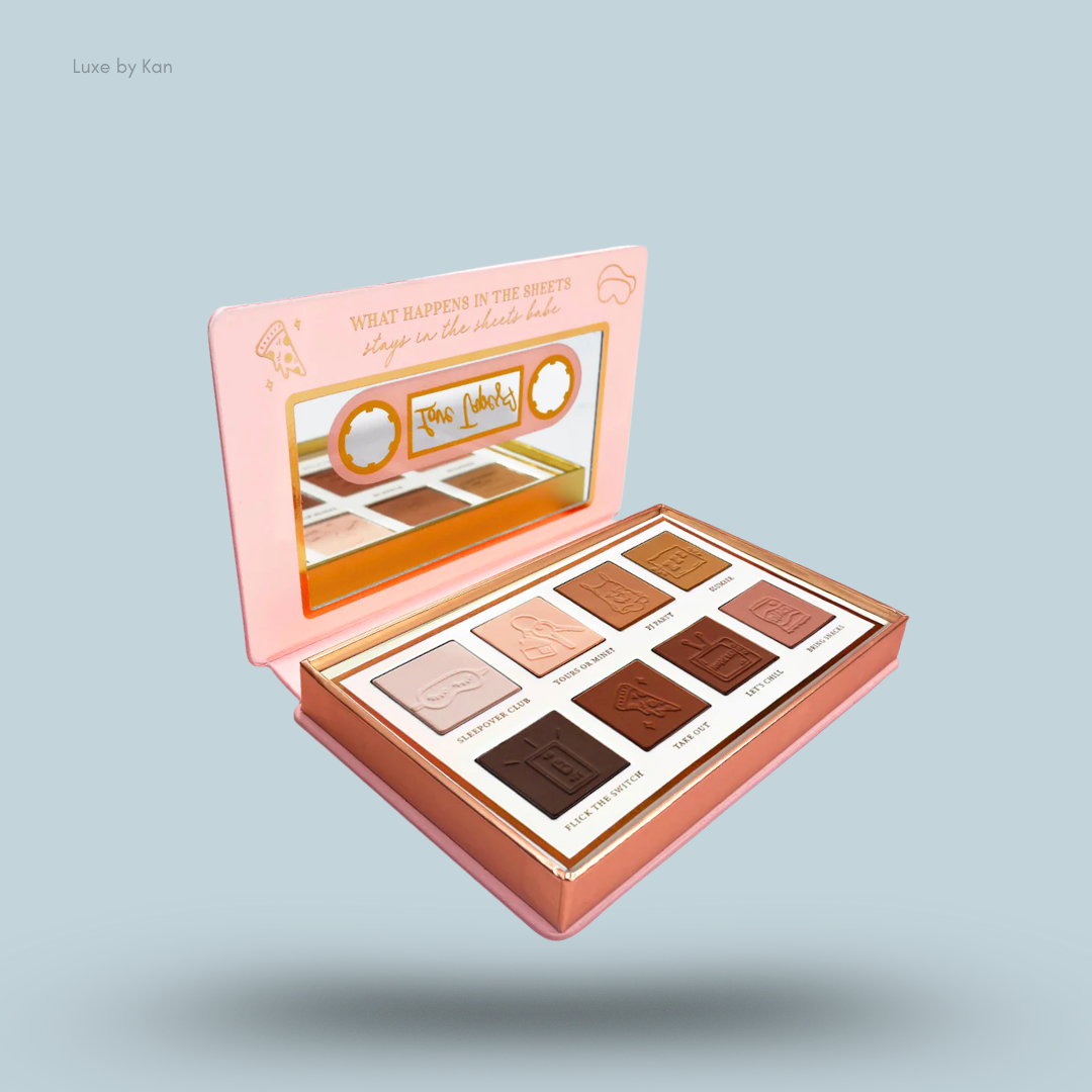 Open eyeshadow palette with pink packaging resembling a cassette tape and containing ten square-shaped eyeshadows in neutral and brown tones, each with a unique debossed image.