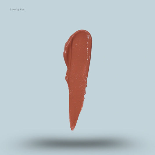 A glossy, warm terracotta lipstick smear floats against a light blue background, showcasing its rich color and smooth texture.