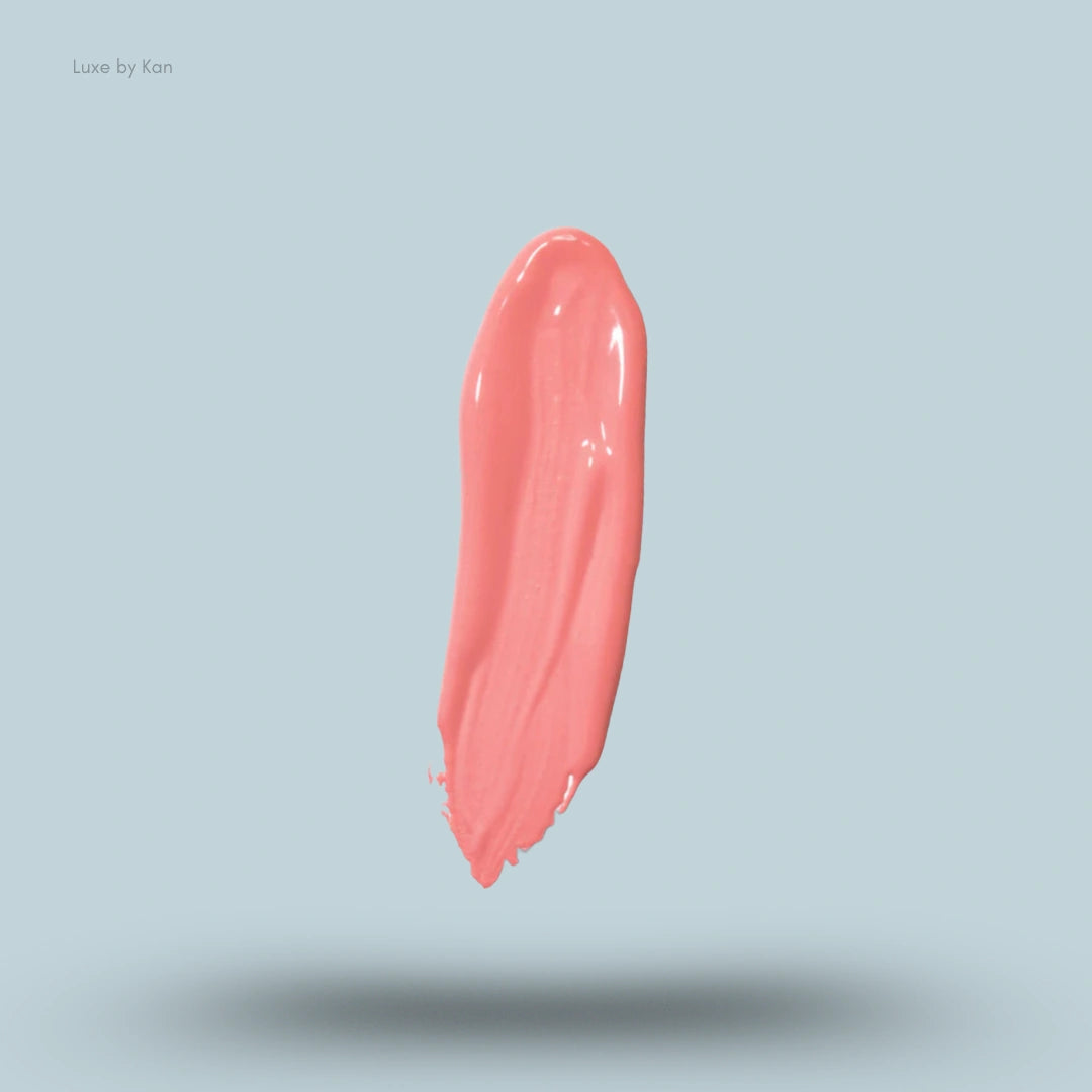 A swatch of Plouise "the cheek of it" liquid blush, shown against a light blue background. The swatch is of an opaque, coral-colored liquid blush.