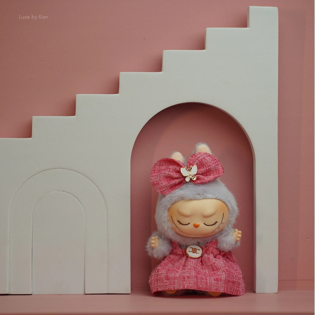 Labubu doll in a pink tweed dress with a matching bow against a pink wall with white stairs.